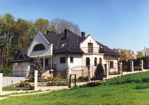 Duży, biały dom jednorodzinny z ciemnym dachem, balkonem z kutą balustradą i ogrodzeniem z kamiennymi słupkami, otoczony zielonym trawnikiem i drzewami.