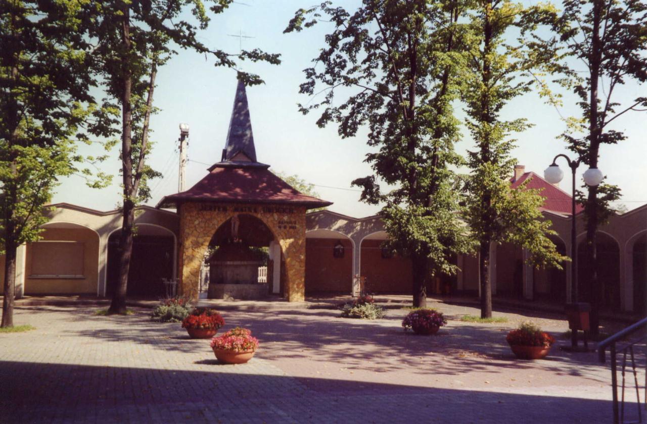 Kamienna altana z wieżyczką, otoczona chodnikiem z kostki brukowej i donicami z kwiatami, na tle budynków z łukowymi podcieniami.