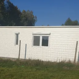 Biały budynek z panelami elewacyjnymi i roletami zewnętrznymi, widziany z niskiej perspektywy, z widoczną siatką ogrodzeniową i zieloną trawą.