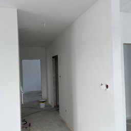 ALFA TAR Robert Tarnowski - Białe, świeżo pomalowane ściany wewnątrz budynku, widoczne puszki z farbą i narzędzia malarskie. Surowe wykończenie, widoczne niedoskonałości i detale. Jasne, naturalne światło.