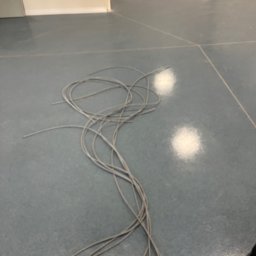 ALFA TAR Robert Tarnowski - Szare przewody ułożone na szarej, błyszczącej podłodze z widocznymi fugami. W tle fragment ściany i sprzęt budowlany. Ujęcie z góry, z bliska.