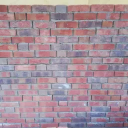 Ściana z czerwonej cegły klinkierowej, z widocznymi spoinami wypełnionymi szarą zaprawą, z fragmentem niebieskiego ubrania w prawym dolnym rogu.