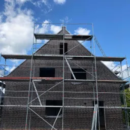 Murowany dom w trakcie budowy, widoczne rusztowania, ściany z ciemnej cegły, otwory okienne i drzwiowe, błękitne niebo z chmurami w tle.