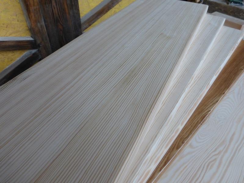 Jasne deski z wyraźnym usłojeniem, ułożone warstwowo. Widoczny fragment konstrukcji drewnianej w tle. Detal drewna.