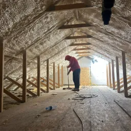 Poddasze ocieplone pianką PUR, widok z perspektywy osoby stojącej na podłodze, pracownik w czerwonej bluzie wygładza pianę na suficie za pomocą narzędzia na długim kiju, w tle światło dzienne...