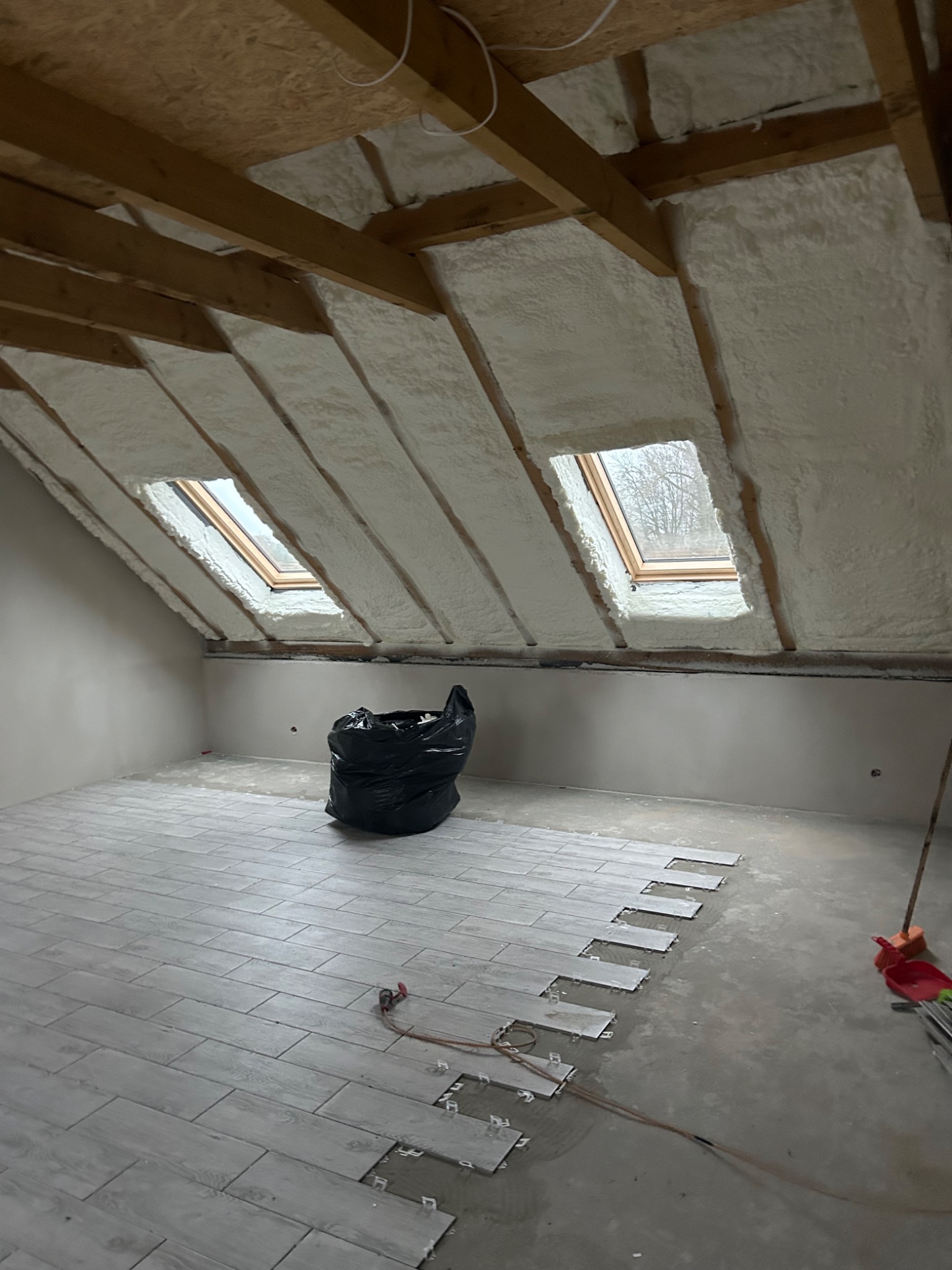 Poddasze z oknami dachowymi po ociepleniu pianką PUR, widoczne drewniane krokwie, częściowo ułożona podłoga z płytek imitujących drewno, czarny worek na śmieci w tle.