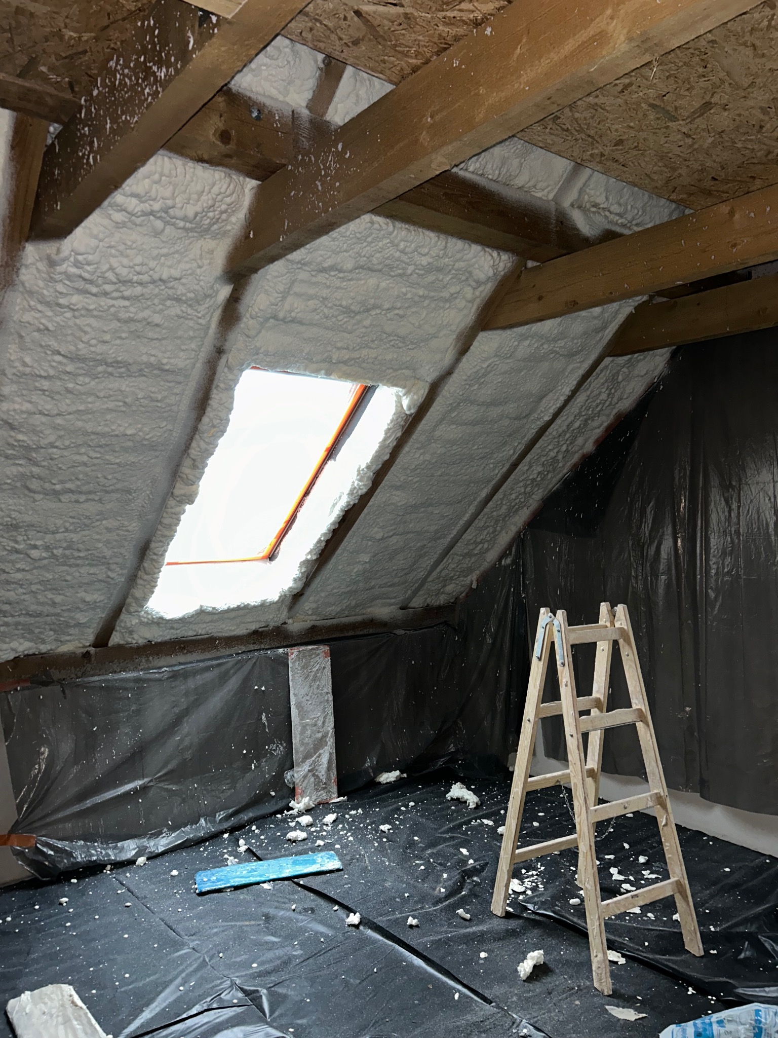 Poddasze ocieplone pianką PUR z widocznym oknem dachowym i drewnianą drabiną na czarnej folii ochronnej.