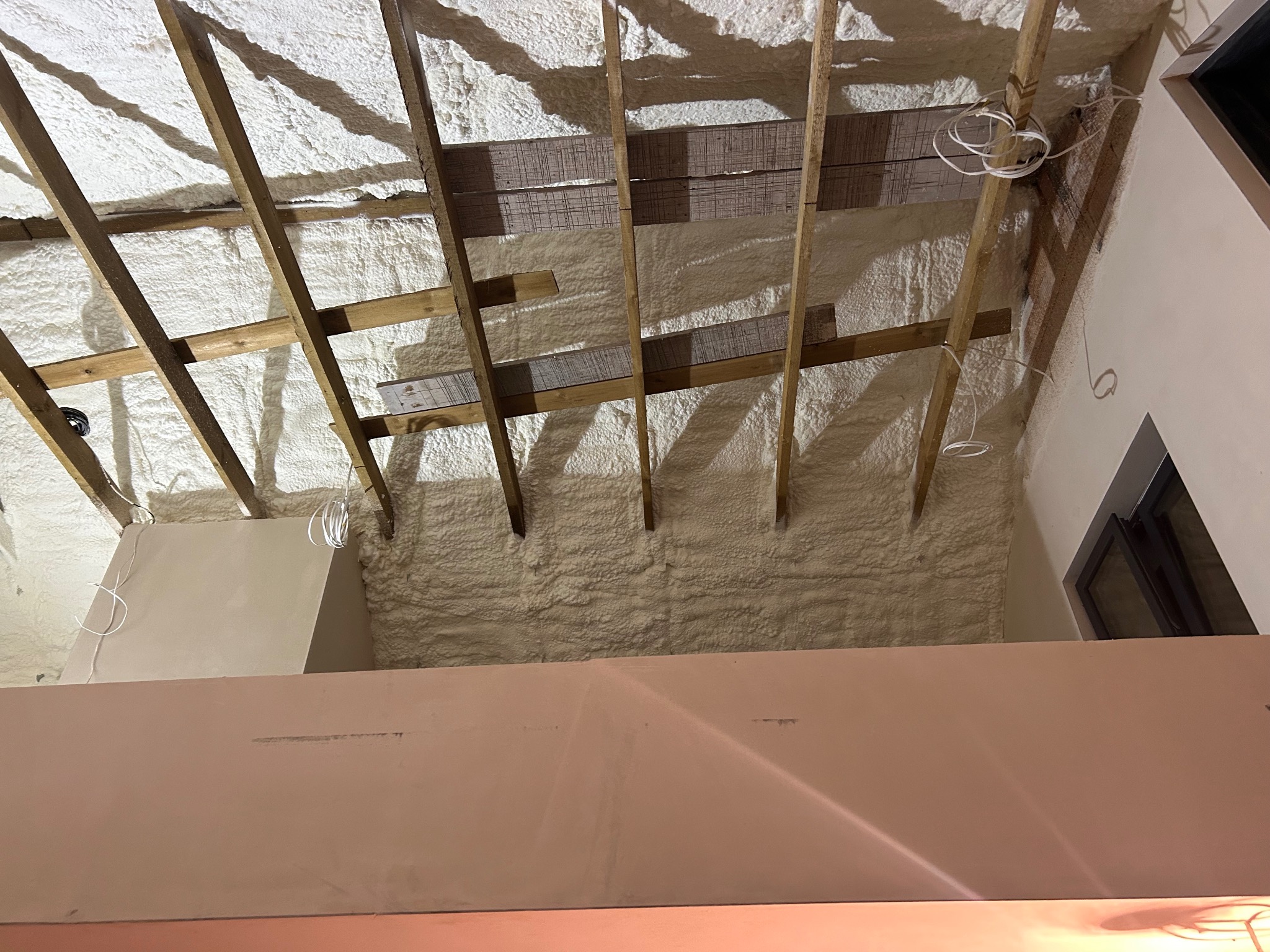 Poddasze z drewnianą konstrukcją dachu pokryte warstwą jasnej pianki izolacyjnej, widoczne przewody elektryczne i fragment ściany z oknem.