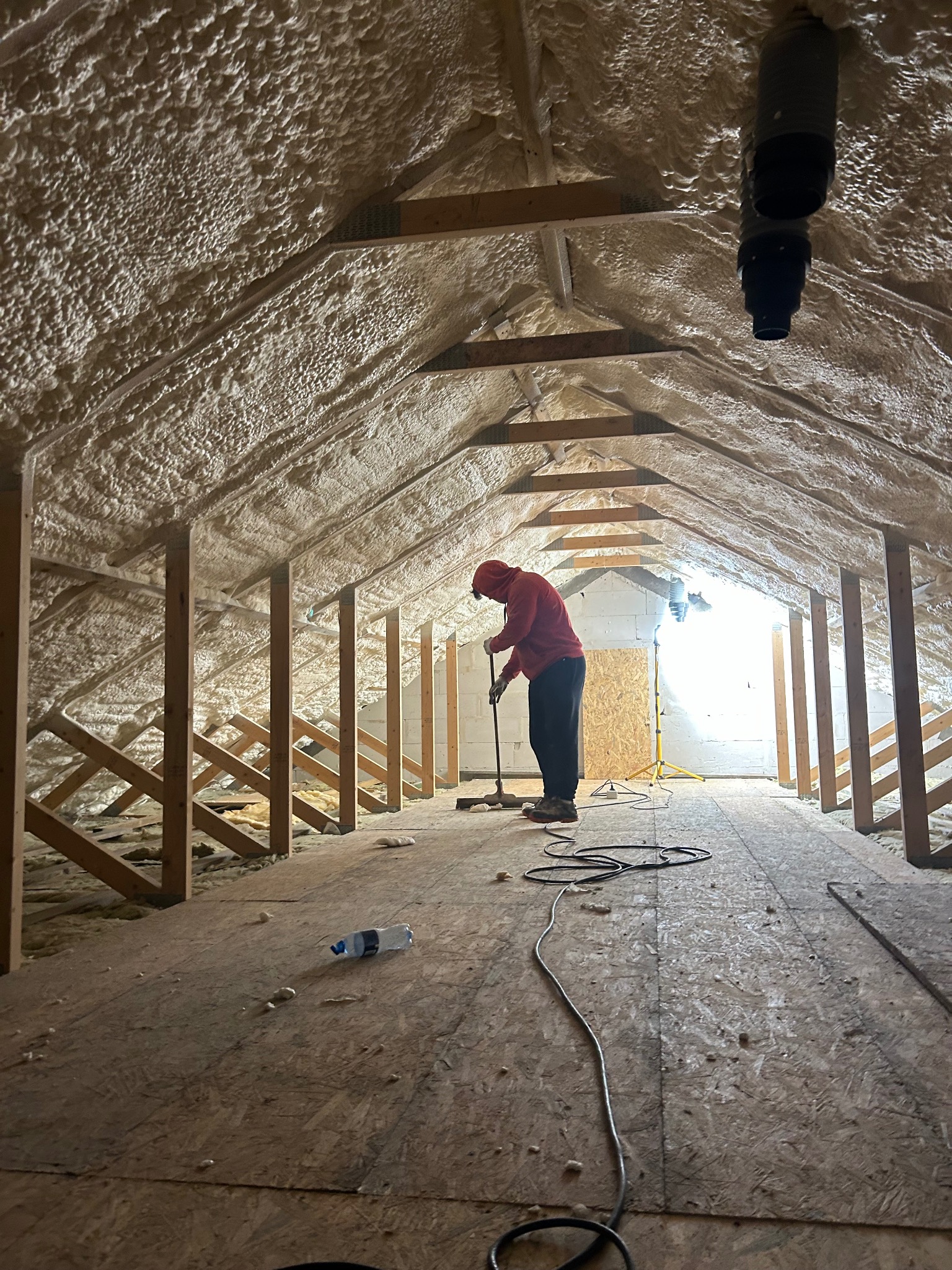 Poddasze ocieplone pianką PUR, widok z perspektywy osoby stojącej na podłodze, pracownik w czerwonej bluzie wygładza pianę na suficie za pomocą narzędzia na długim kiju, w tle światło dzienne...