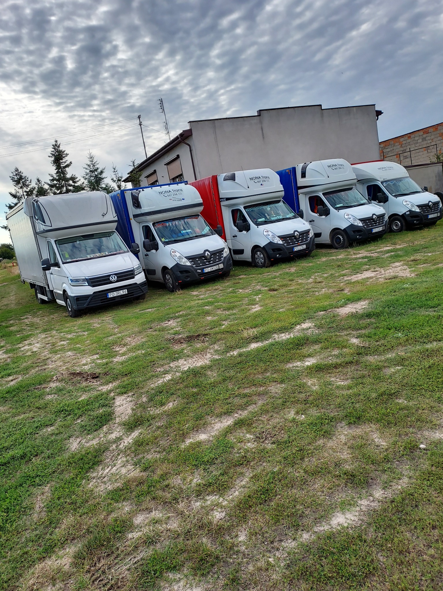 Flota pięciu białych busów dostawczych zaparkowana na trawiastym terenie przed budynkiem w pochmurny dzień, widoczne numery rejestracyjne i logo firmy.