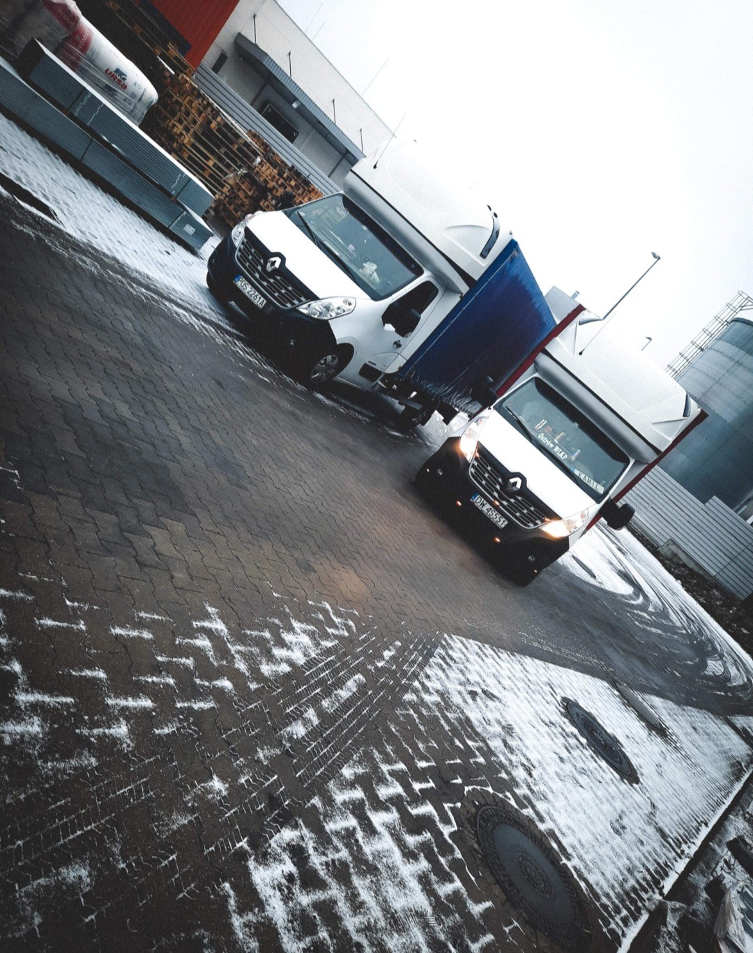 Dwa białe busy Renault Master z plandekami na mokrej, częściowo ośnieżonej kostce brukowej na placu manewrowym. W tle widoczne magazyny i silosy.