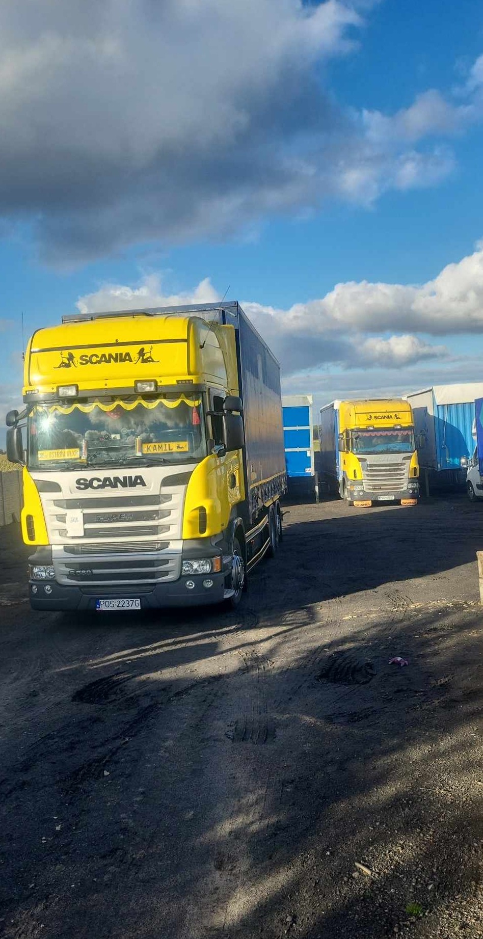 Żółta ciężarówka Scania z imieniem 'Kamil' na przedniej szybie, zaparkowana na placu z innymi ciężarówkami w tle, pod błękitnym niebem z chmurami.