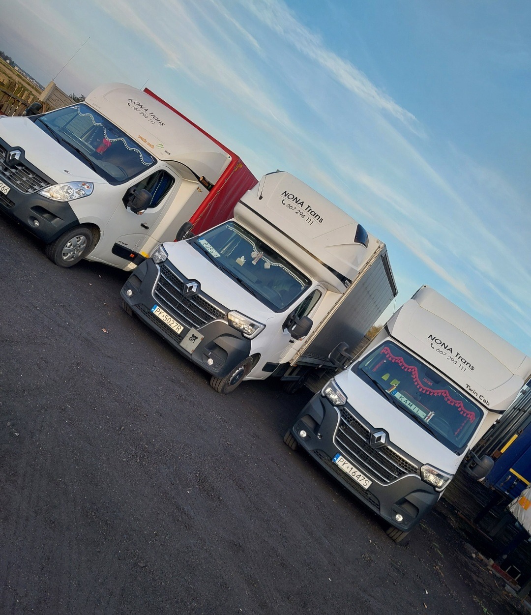 Trzy białe busy transportowe marki Renault ustawione ukośnie na parkingu, z logo firmy 'NONA Trans' widocznym na każdym z nich, z dekoracjami w kabinach i niebieskim niebem w tle.