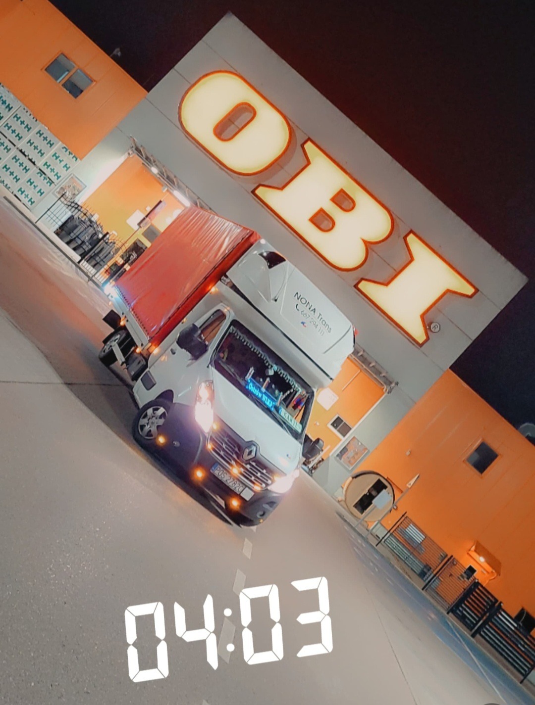 Biały bus z czerwoną plandeką zaparkowany przed sklepem OBI w nocy, widoczny numer rejestracyjny i nazwa firmy transportowej Nona Trans.