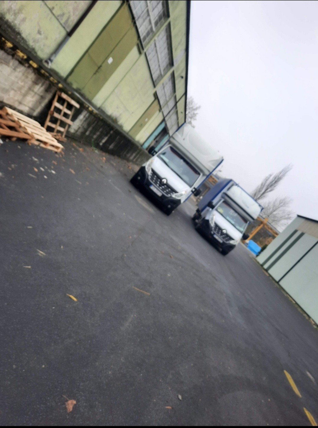 Dwa busy dostawcze, biały i ciemny, zaparkowane na placu przed magazynem w pochmurny dzień. W tle widoczne palety.