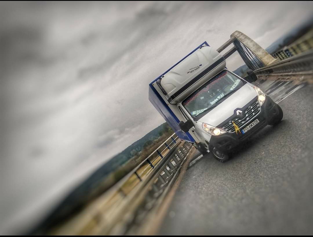 Biały bus Renault z zabudową na dachu, jadący po moście w pochmurny dzień, widoczny z boku pod kątem.