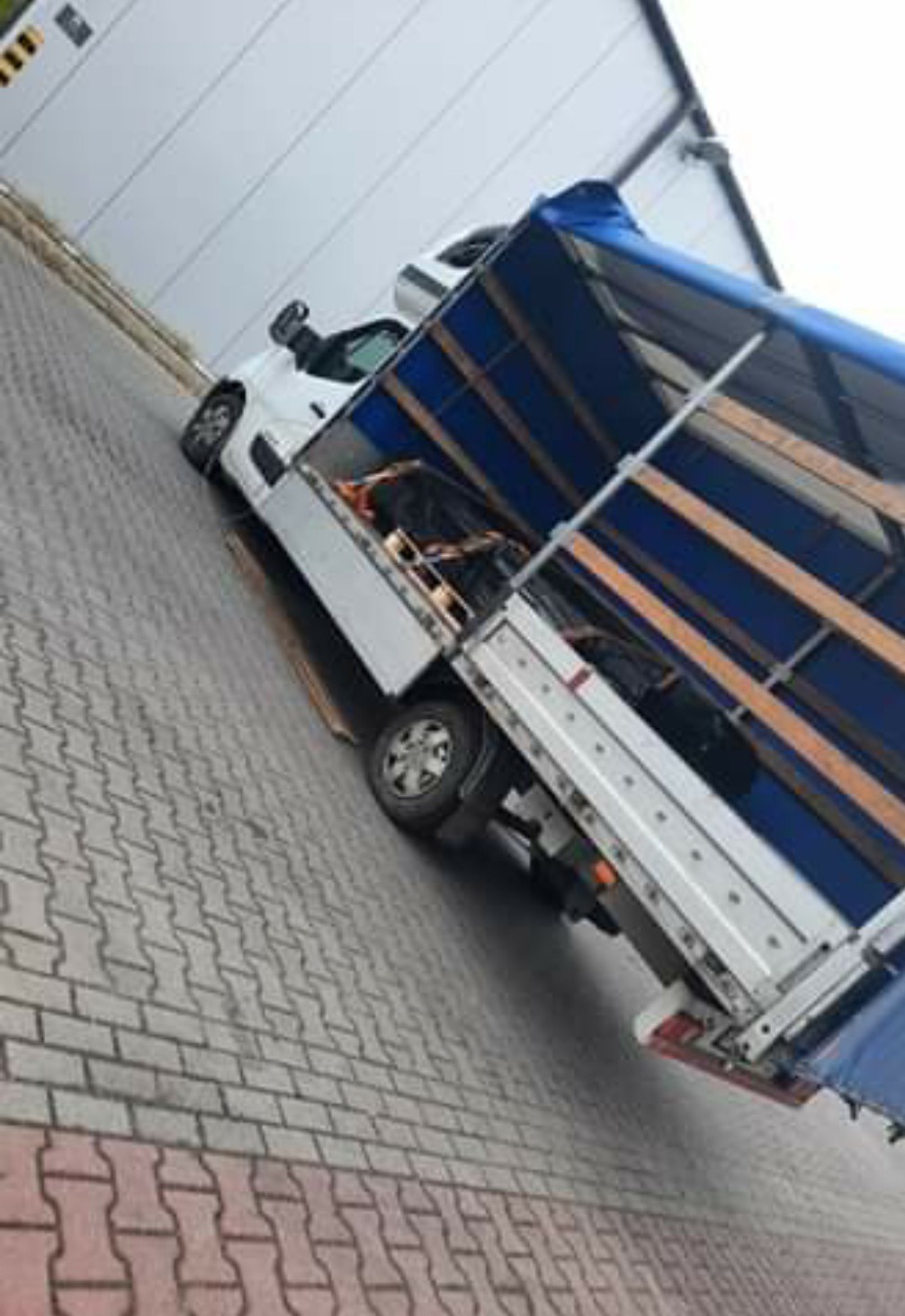 Biały samochód dostawczy z plandeką w kolorze niebieskim, częściowo załadowany towarem na palecie, zaparkowany na chodniku z kostki brukowej przed budynkiem.