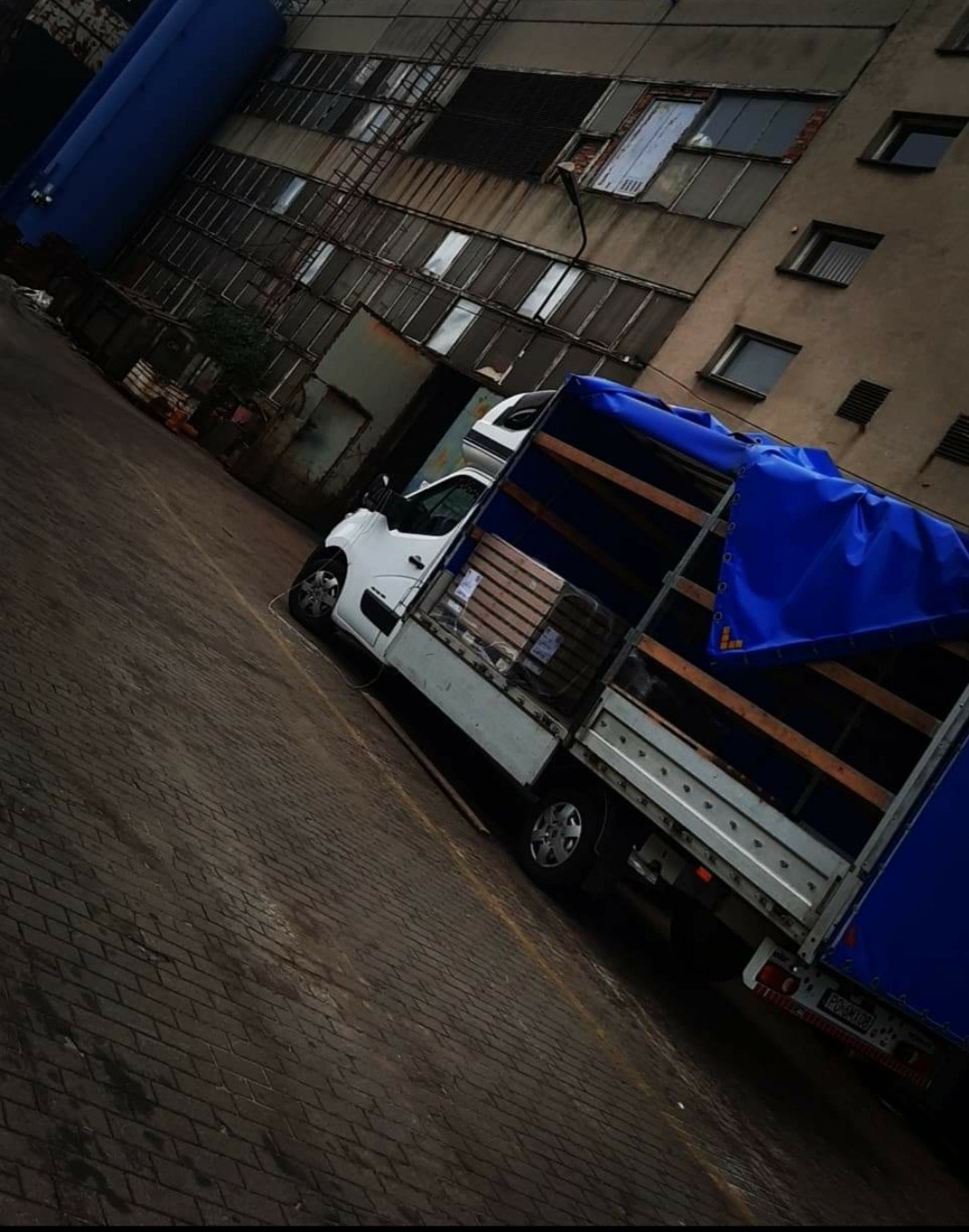 Biały samochód dostawczy z niebieską plandeką zaparkowany na brukowanym placu, z ładunkiem palet wewnątrz, na tle starego, wielopiętrowego budynku przemysłowego.
