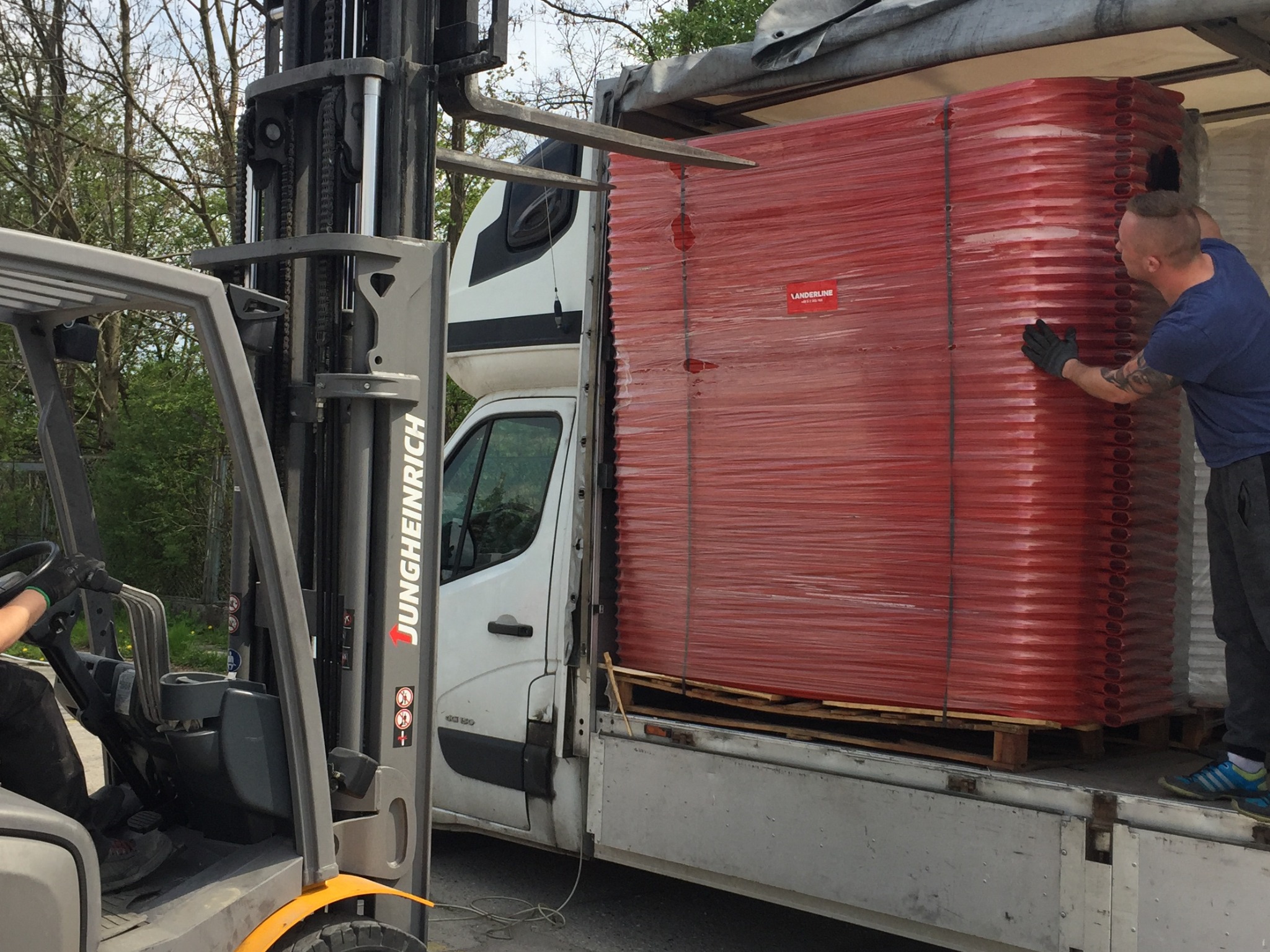 Załadunek czerwonych elementów konstrukcyjnych na palecie do białego busa za pomocą wózka widłowego Jungheinrich, pracownik w rękawicach zabezpiecza ładunek.