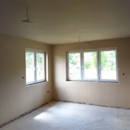 Pokój po nałożeniu gładzi gipsowej na ścianach i suficie, widoczne dwa okna z białymi ramami, surowa podłoga, przygotowane wyprowadzenia elektryczne.