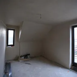 Pokój na poddaszu w trakcie prac wykończeniowych. Ściany i sufit po tynkowaniu, widoczne puszki elektryczne, okno i drzwi balkonowe w czarnych ramach.