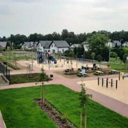 Widok z góry na nowy, uporządkowany plac zabaw z piaskownicą, huśtawkami i zjeżdżalniami, otoczony świeżo posadzoną trawą i drzewami, z widocznymi domami w tle i zaparkowanymi samochodami dostawczymi.