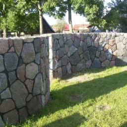 Kamienny murek ogrodowy z nieregularnych, różnokolorowych otoczaków, spoinowany ciemną fugą, na tle zielonego trawnika i zabudowań wiejskich.