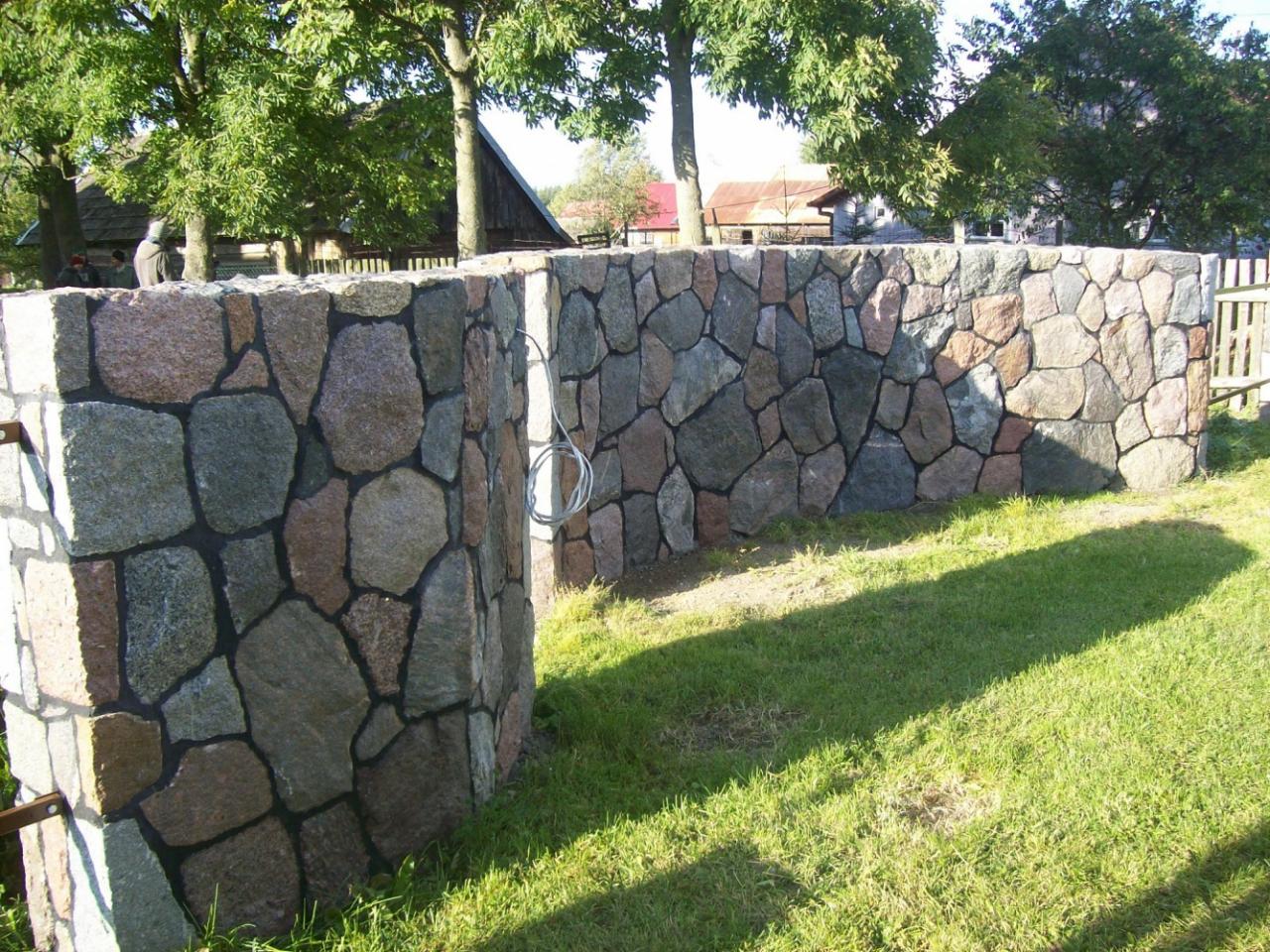 Kamienny murek ogrodowy z nieregularnych, różnokolorowych otoczaków, spoinowany ciemną fugą, na tle zielonego trawnika i zabudowań wiejskich.