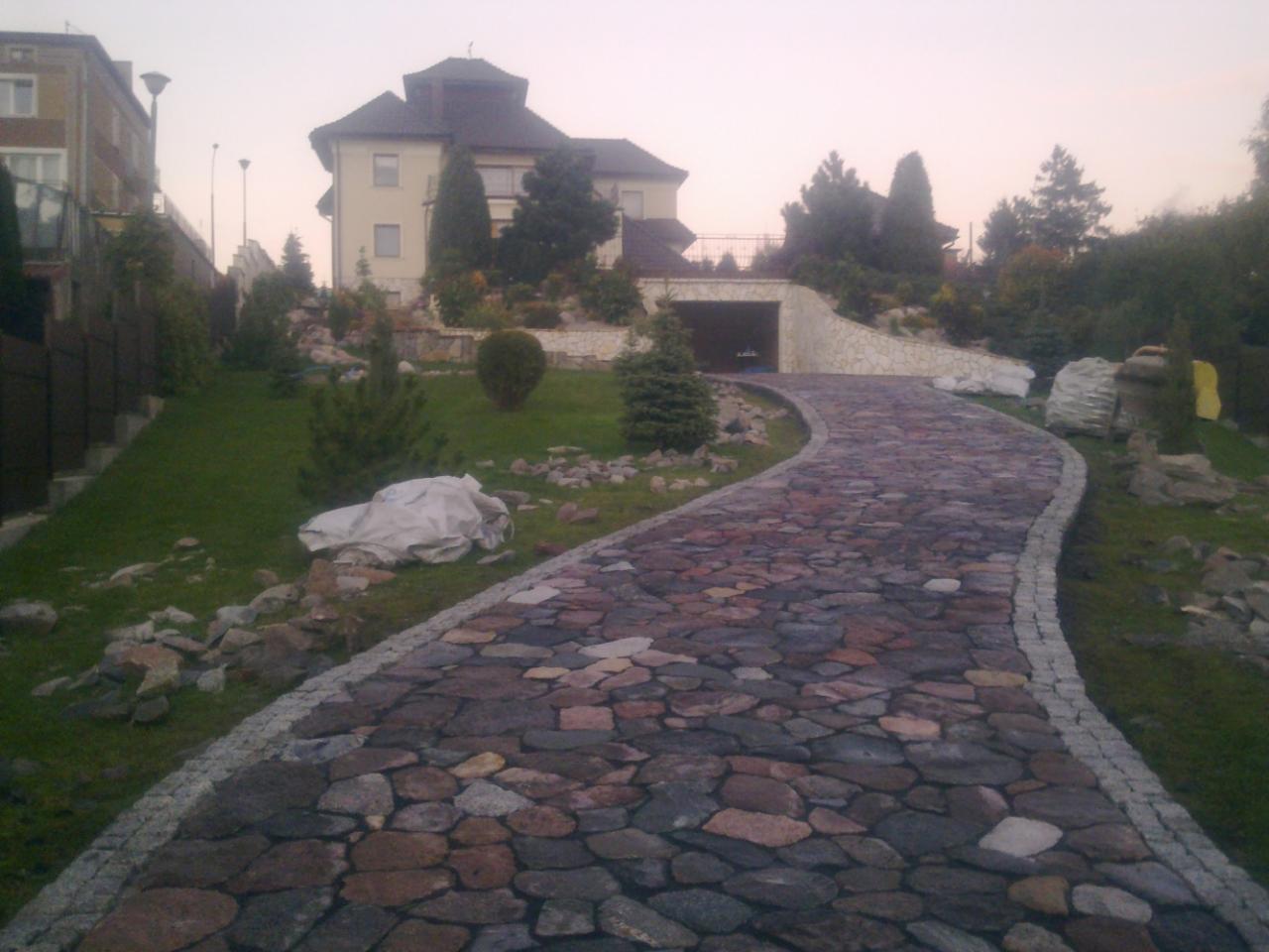 Kręta ścieżka z nieregularnej kostki brukowej prowadząca do garażu wkomponowanego w skarpę, otoczona zielonym trawnikiem i ozdobnymi krzewami, z widocznym budynkiem mieszkalnym w tle.