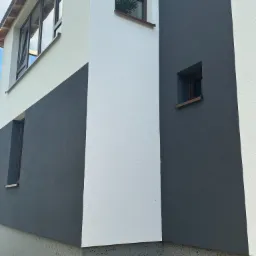 Fragment elewacji budynku z oknami, pomalowanej w dwóch odcieniach szarości i bieli, z widoczną fakturą tynku.