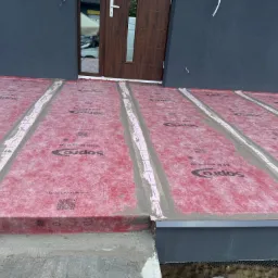 Przygotowanie do hydroizolacji tarasu z użyciem membrany Sopro AEB Plus 639, widoczne pasy uszczelniające na łączeniach, szara elewacja budynku w tle.