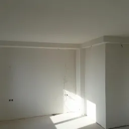 Wykończone wnętrze w stanie deweloperskim: białe ściany, gładzie, listwy przysufitowe, wystające kable elektryczne, butelka wody w rogu.