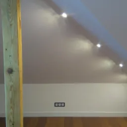 Wykończone poddasze ze skośnym sufitem, wbudowanymi punktowymi lampami LED, drewnianą kolumną konstrukcyjną i trzema gniazdkami elektrycznymi w czarnej ramce na ścianie.