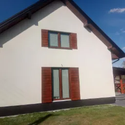 Świeżo otynkowana, biała fasada domu jednorodzinnego z drewnianymi okiennicami w kolorze orzechowym, widok z poziomu trawnika w słoneczny dzień.