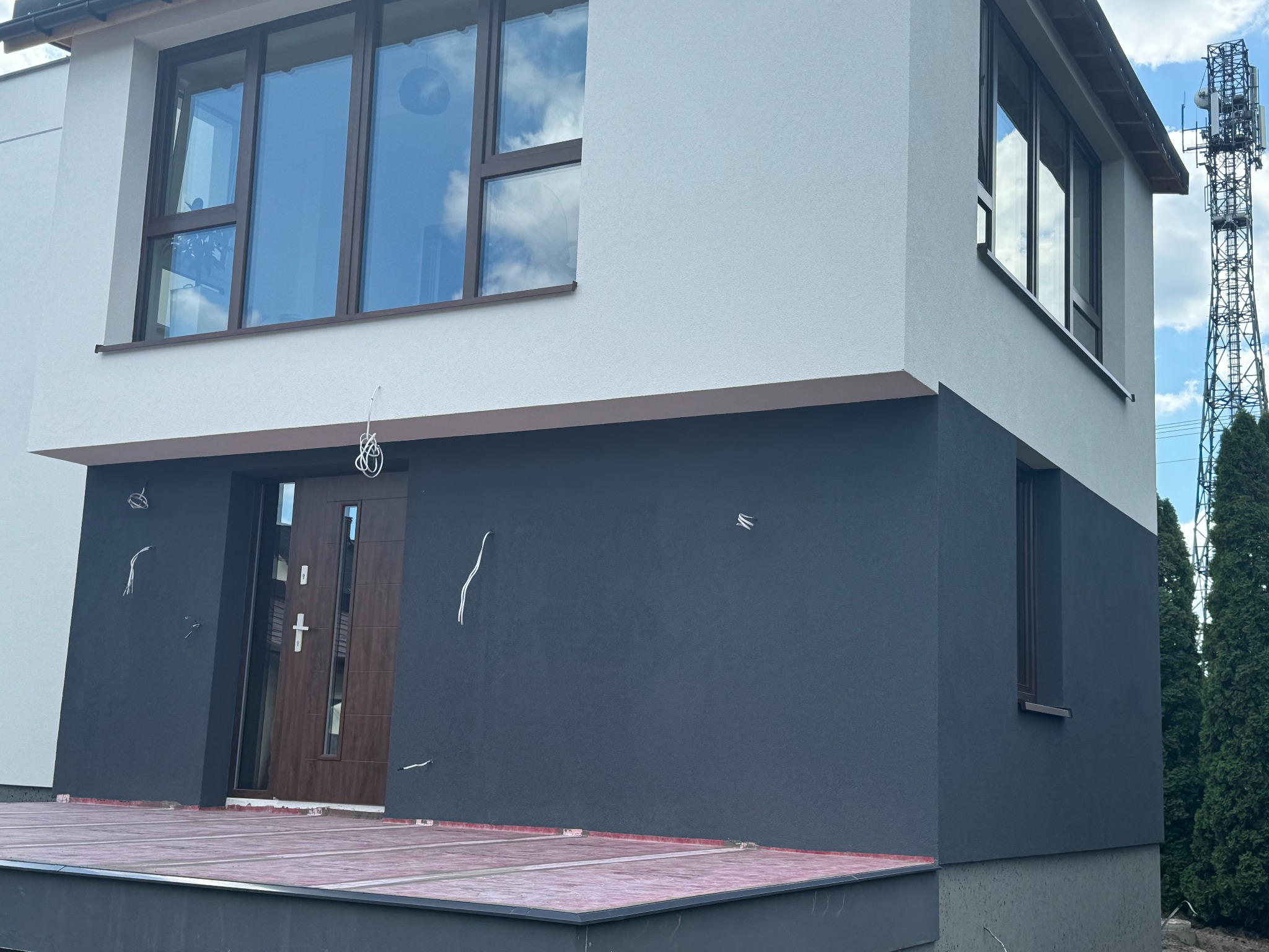 Nowoczesny dom dwukondygnacyjny z elewacją w kolorze białym i grafitowym, widoczne wystające przewody elektryczne na ścianie, brązowe okna i drzwi wejściowe, w tle maszt telekomunikacyjny.