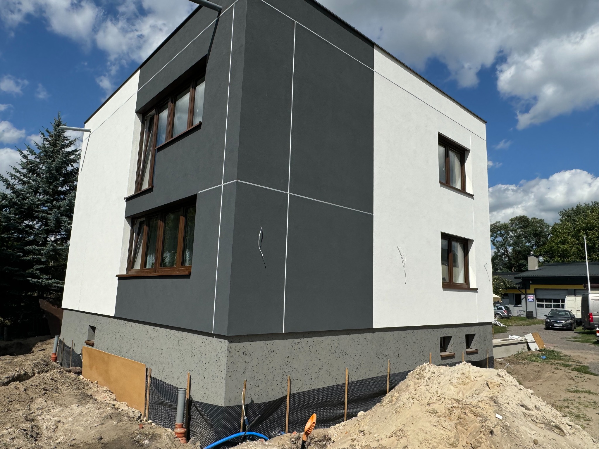 Nowoczesny dom jednorodzinny z elewacją w kolorach bieli i grafitu, widoczne prace ziemne wokół fundamentów.