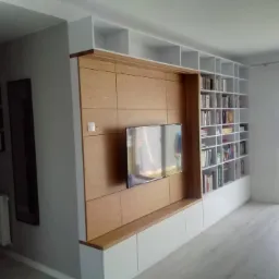 Nowoczesna zabudowa ściany w salonie: panele z jasnego drewna z wbudowanym telewizorem, białe szafki dolne i regał na książki z białymi półkami, całość podświetlona naturalnym światłem wpadającym...