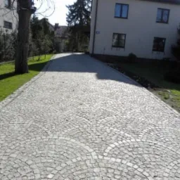 Ułożona z szarej kostki granitowej droga do domu, wzór wachlarza. Widok na budynek i zieleń po bokach. Kąt ujęcia z dołu, podkreślający wzór.