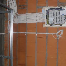 Rozdzielnica elektryczna w stanie surowym, widoczne przewody prowadzone po ścianie z cegły oraz metalowa konstrukcja wsporcza.