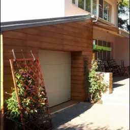 Nowoczesny dom z garażem obłożonym drewnianą boazerią, z kratką na pnącza opartą o ścianę i roślinami w doniczce, widoczny fragment kostki brukowej i meble ogrodowe w tle.