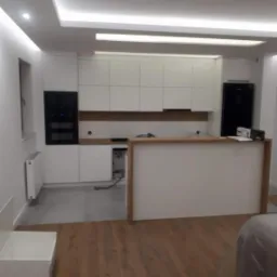 Wnętrze kuchni w trakcie montażu: białe szafki kuchenne z czarnym piekarnikiem, drewniany blat, szara podłoga, widoczne kable i niedokończone podłączenia.