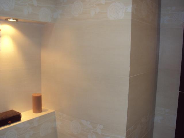 Beżowe płytki z delikatnym motywem różanym zdobią ścianę łazienki, oświetlona półka z brązowym pudełkiem i świecą w rogu.