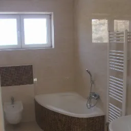 Wykończona łazienka z beżowymi płytkami imitującymi marmur, narożna wanna obudowana mozaiką, bidet, grzejnik drabinkowy i okno z widokiem na zewnątrz.