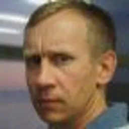 Portret mężczyzny o krótkich włosach, zamyślony wyraz twarzy, w niebieskiej koszuli, na rozmytym tle z elementami różu i szarości.