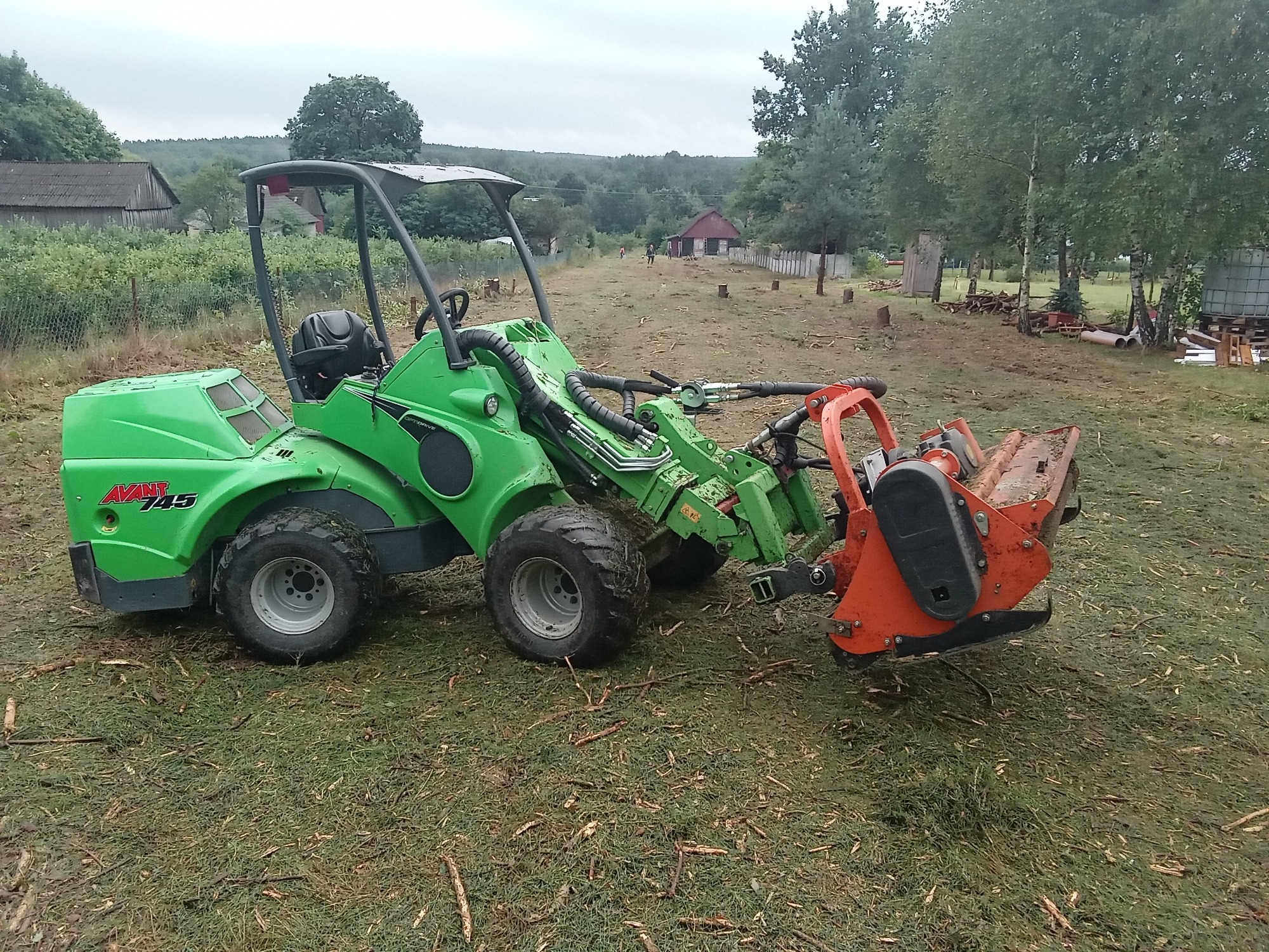 Zielona maszyna Avant 745 z pomarańczowym frezem na trawniku z resztkami gałęzi, w tle budynki gospodarcze i drzewa.