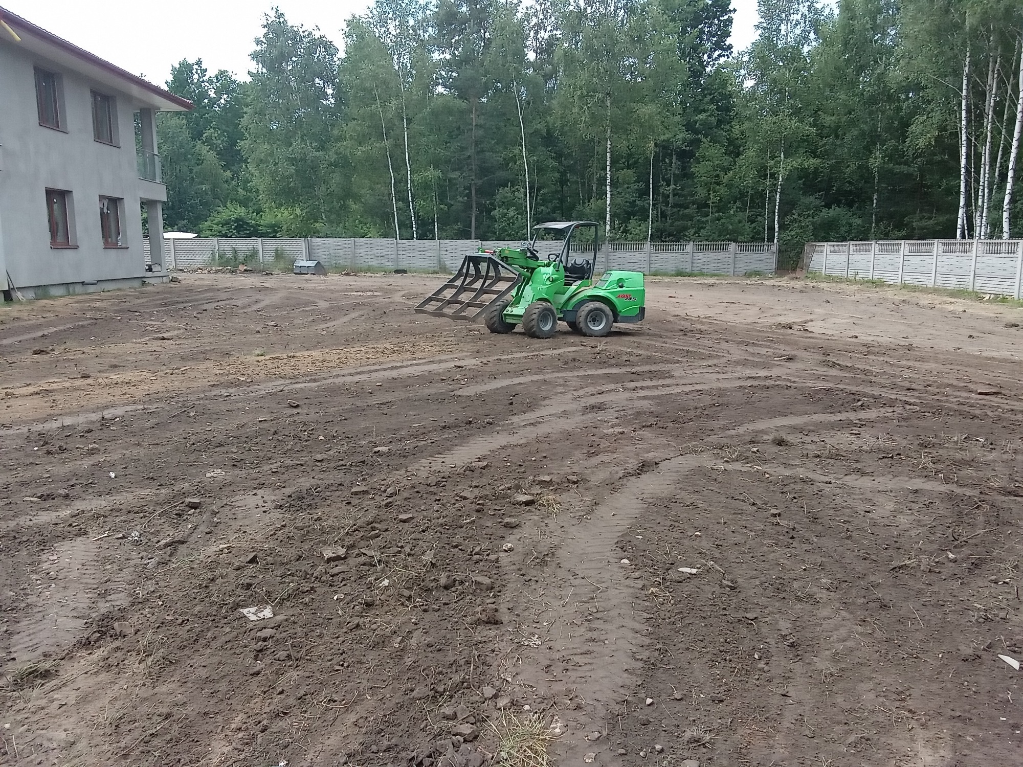 Zielony miniładowarka Avant 750 z osprzętem do wyrównywania terenu na przygotowanym pod trawnik placu budowy, widoczne ślady opon na ubitej ziemi, w tle budynek w stanie surowym i betonowe ogrodzenie.