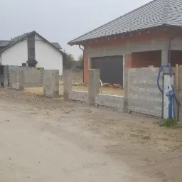 Budowa domu jednorodzinnego: widok na wznoszone ściany z bloczk&oacute;w betonowych, z widocznymi workami zaprawy i taczką na placu budowy, na tle innych dom&oacute;w.