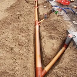Pomarańczowe rury kanalizacyjne ułożone w wykopie, widoczne połączenia i fragmenty studzienek, w tle ogrodzenie i budynek.