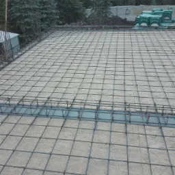 Zbrojenie stropu z siatki stalowej na deskowaniu z płyt OSB, widoczne wystające pręty i elementy konstrukcyjne, w tle drzewa i materiały budowlane