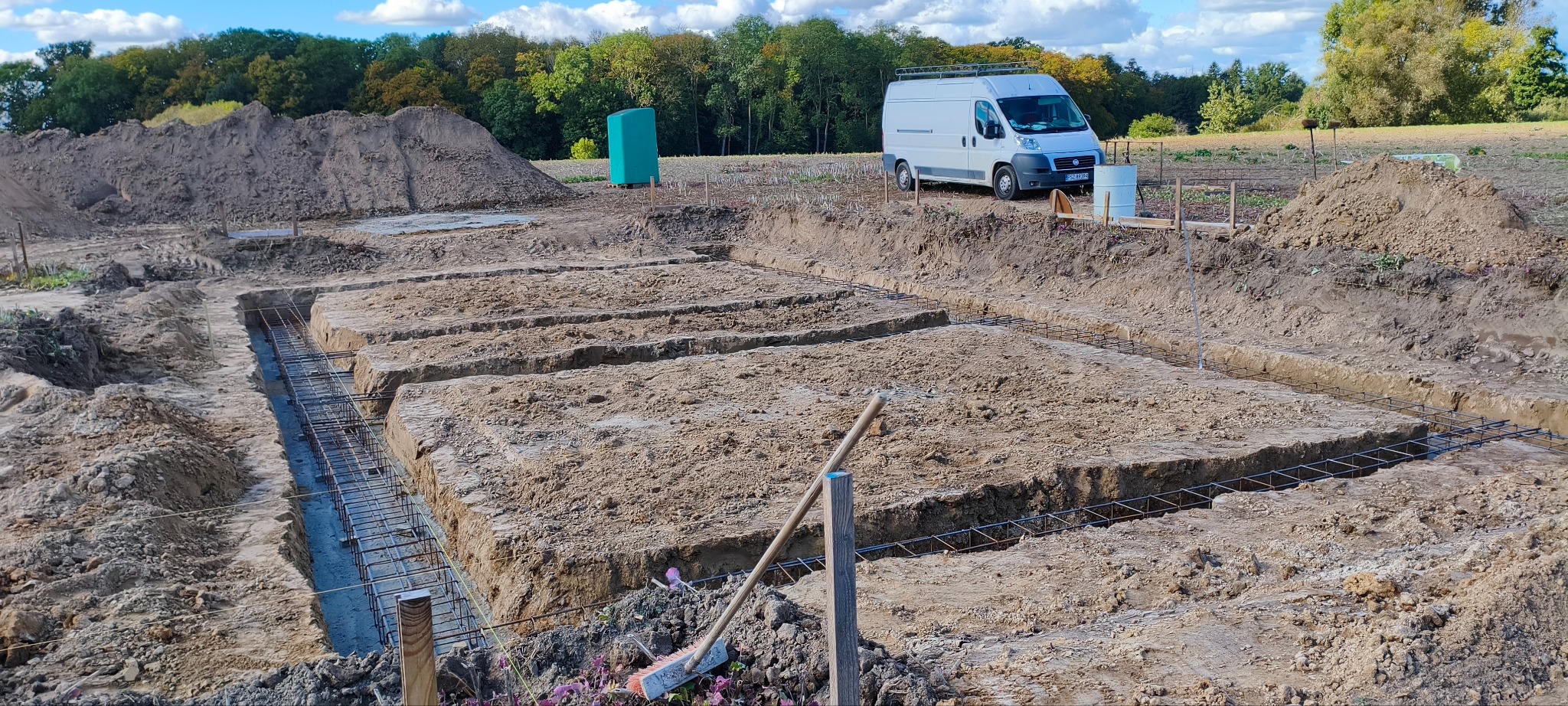 Przygotowanie terenu pod fundamenty: wykopane rowy z częściowo zalanym betonem i zbrojeniem, w tle hałda ziemi, biały bus i zielony zbiornik.